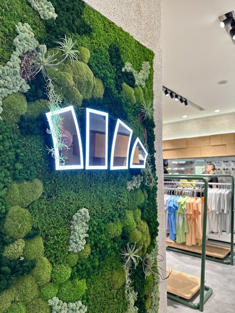 A vibrant green moss wall with a glowing BENEUNDER logo, showcasing the shop's modern interior and apparel.