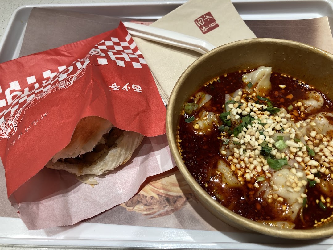 A tray featuring a roujiamo (meat burger) in a red wrapper and a bowl of spicy wontons with peanuts.