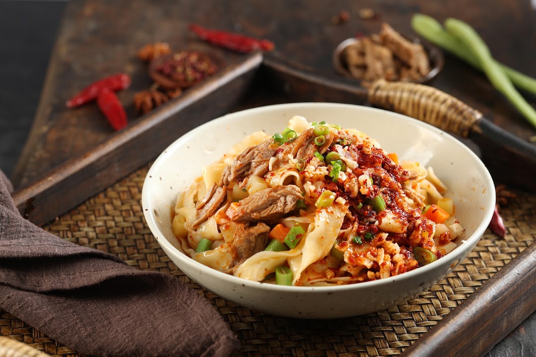 A vibrant bowl of spicy noodles with tender meat and fresh garnishes, served on a woven mat.