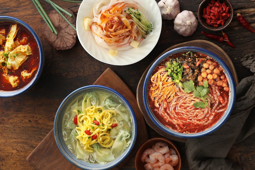 A vibrant overhead view of multiple Chinese noodle and dumpling dishes on a rustic wooden table.
