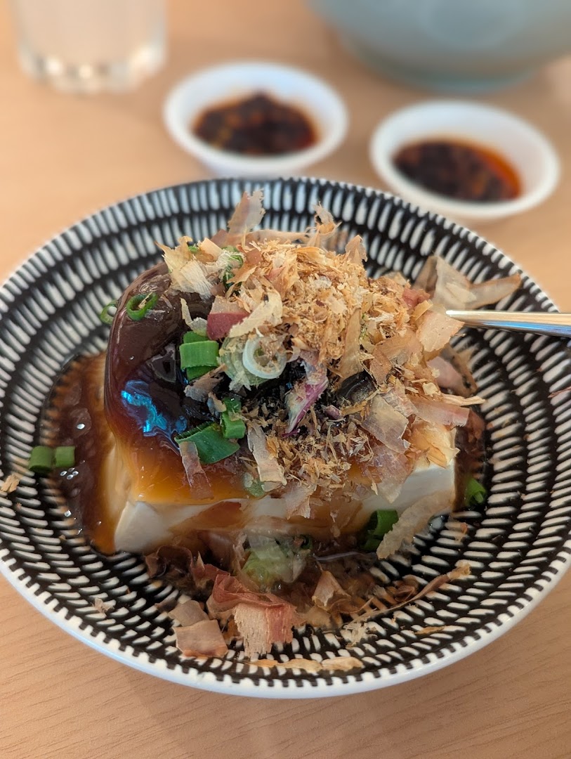 A close-up of a delicious tofu dish topped with bonito flakes, spring onions, and a savory sauce.