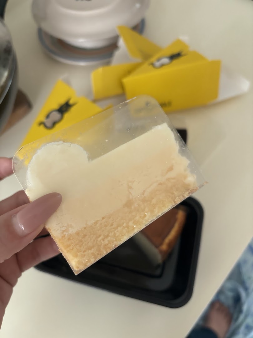 A hand holds a slice of creamy cheesecake, with yellow Cat the Fiddle cake boxes in the background.