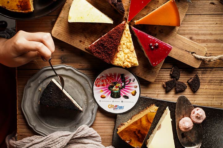 A vibrant assortment of cheesecake slices and desserts on a rustic wooden table, with a hand serving a slice.