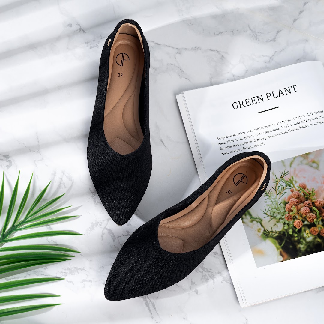 A pair of black pointed-toe flats displayed on a marble surface with a magazine and green foliage.