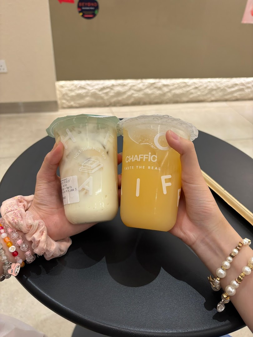Two hands hold refreshing Chaffic beverages, one milk tea and one fruit drink, over a black table.