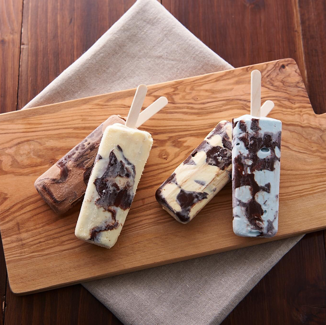Four colorful, swirled ice cream bars on a wooden cutting board, showcasing various delicious flavors.