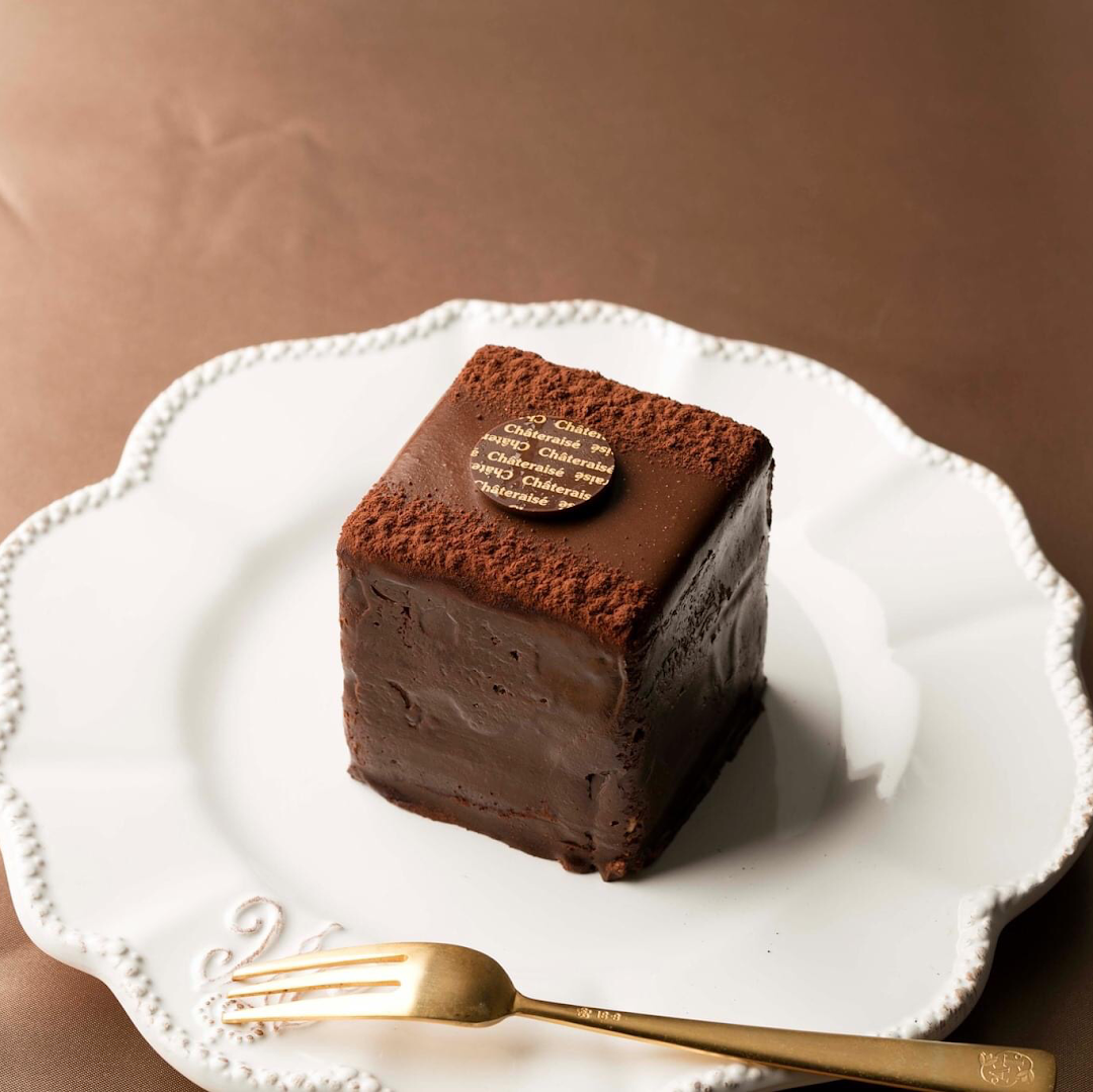 A rich, square chocolate cake with cocoa powder and a Châteraisé logo on a white plate with a gold fork.