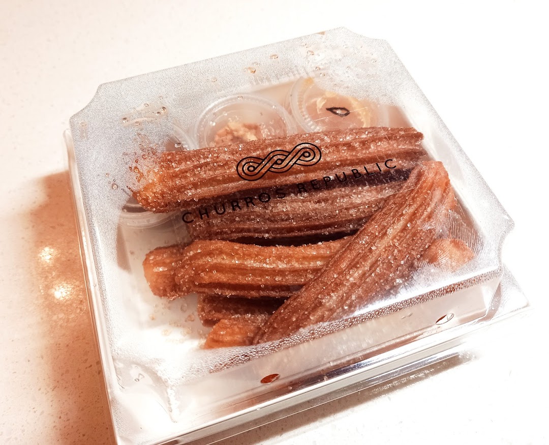 A clear plastic box filled with sugar-dusted churros and small dipping sauce containers from Churros Republic.