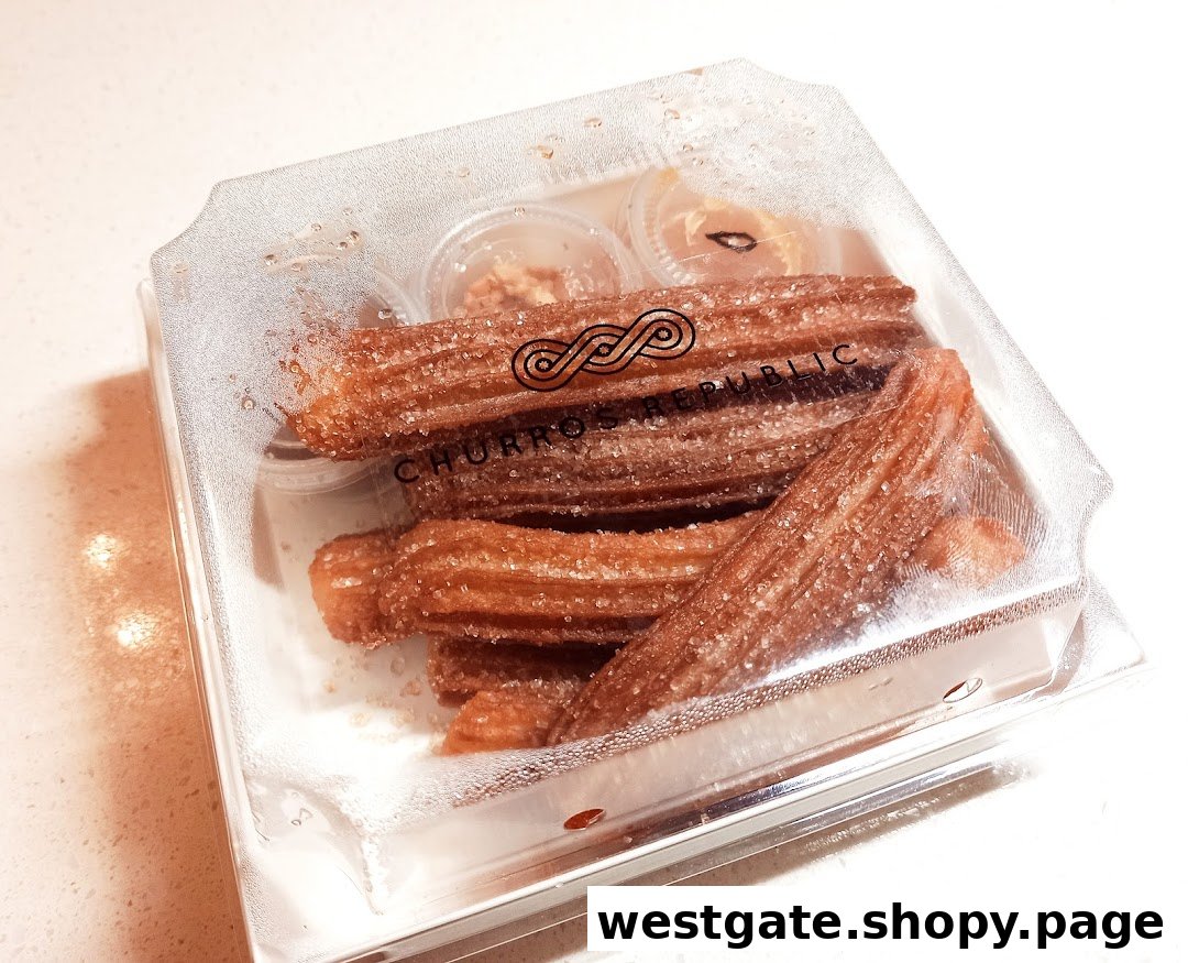A clear plastic box filled with sugar-dusted churros and small dipping sauce containers from Churros Republic.
