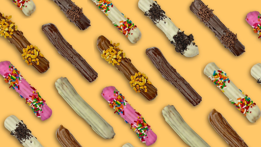 A vibrant pattern of various churros, some coated in chocolate, white chocolate, or pink frosting with sprinkles and nuts.