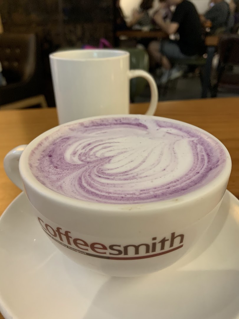 A close-up of a purple latte with latte art in a Coffeesmith cup, with a blurred interior background.