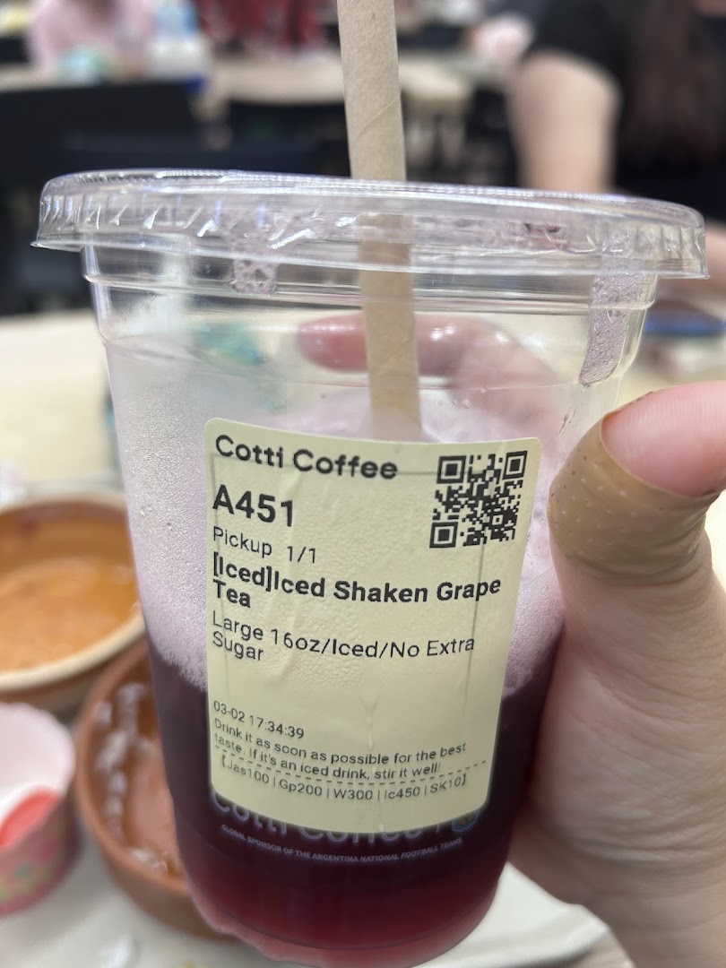 A hand holds a clear cup of Cotti Coffee's Iced Shaken Grape Tea with a label.