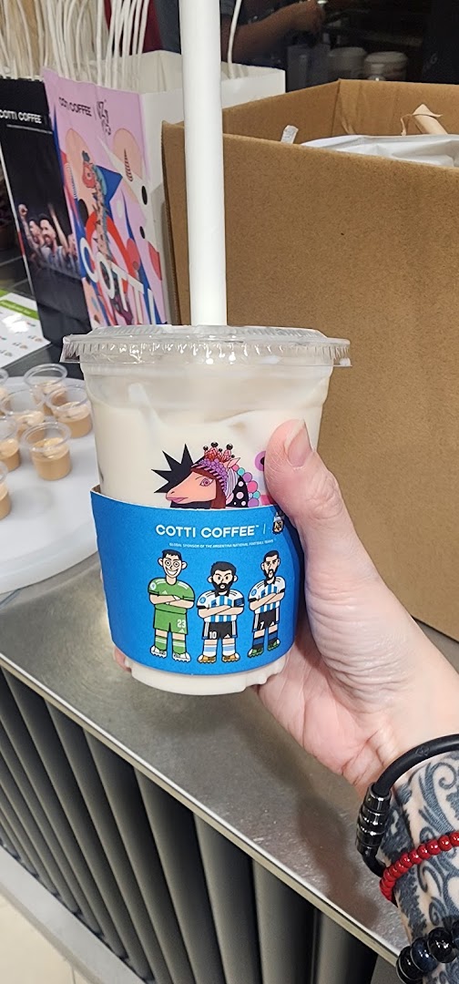 A hand holds a Cotti Coffee drink with a special blue sleeve featuring cartoon football players.