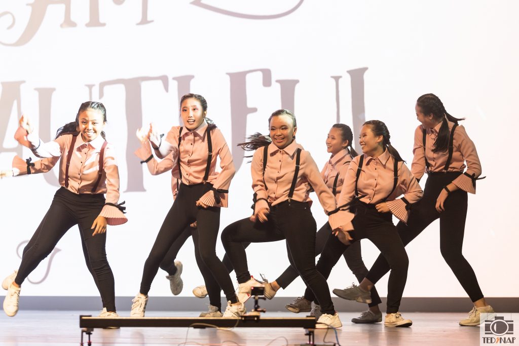 A group of young dancers in pink shirts and black pants performing on stage.