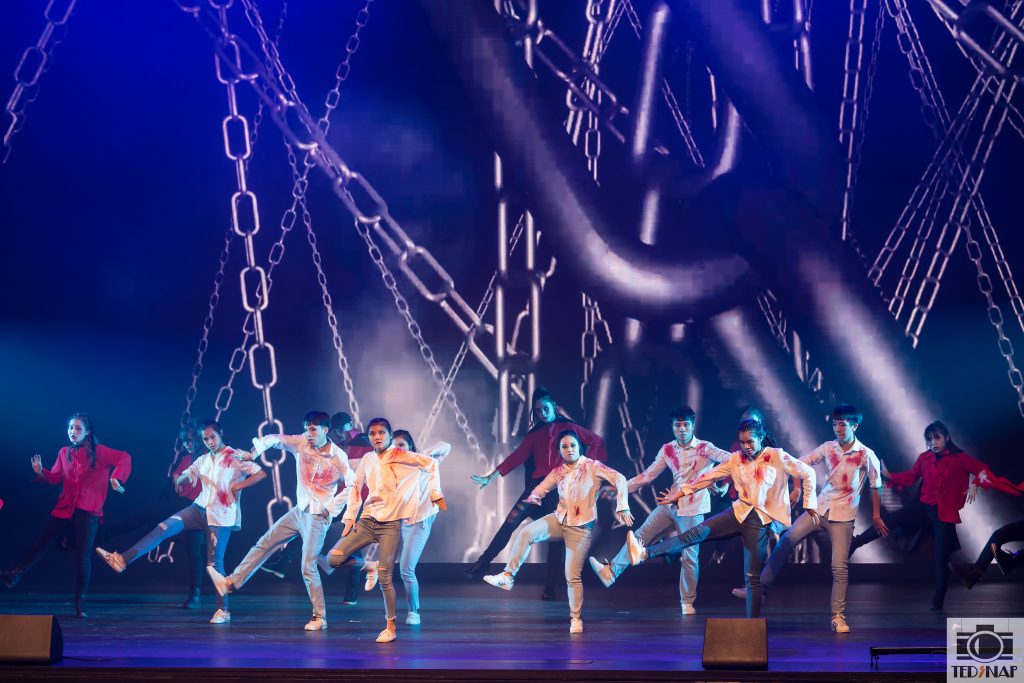 A group of young dancers performs on stage with dramatic lighting and a digital chain backdrop.