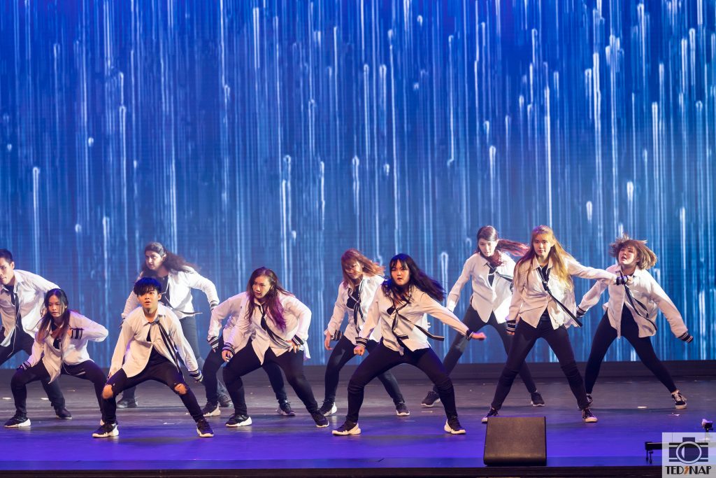 A group of dancers performing on a stage with a dynamic blue light background.