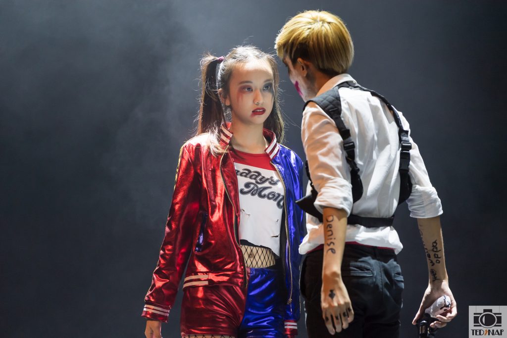 Two individuals in elaborate costumes, one resembling Harley Quinn, on a dimly lit stage.
