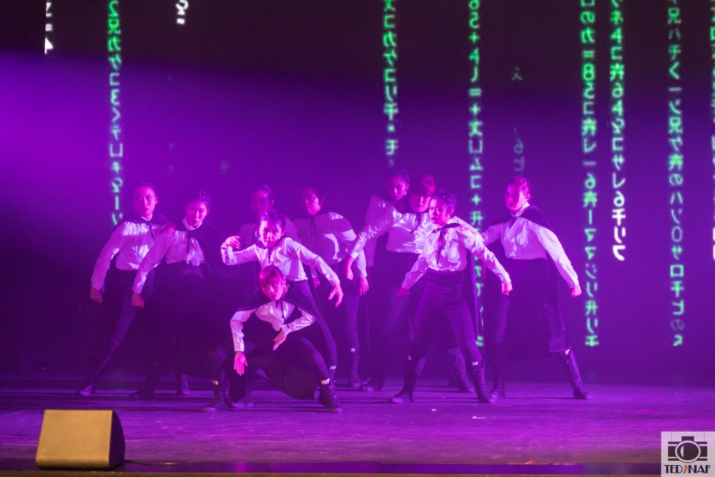 A group of performers in white shirts and black attire on a stage with purple lighting and digital projections.