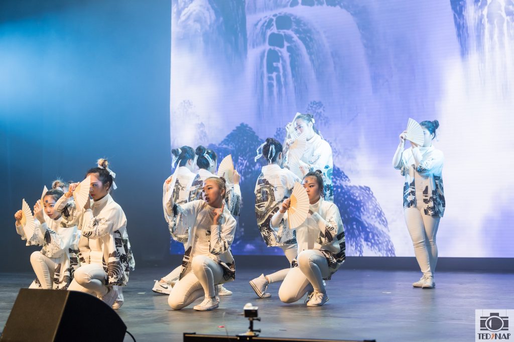 Dancers in white costumes with fans perform on a stage with a scenic backdrop.