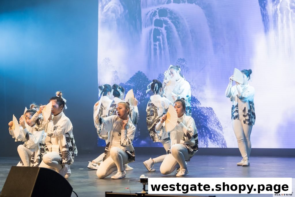 Dancers in white costumes with fans perform on a stage with a scenic backdrop.