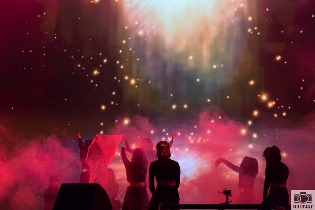 Performers on stage amidst vibrant lights and smoke, showcasing dynamic movement.