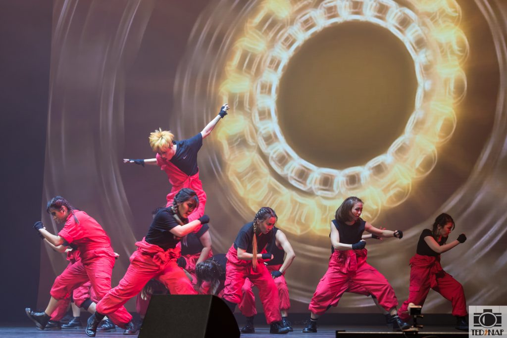A group of dancers in red and black performing on a stage with a dynamic light projection.