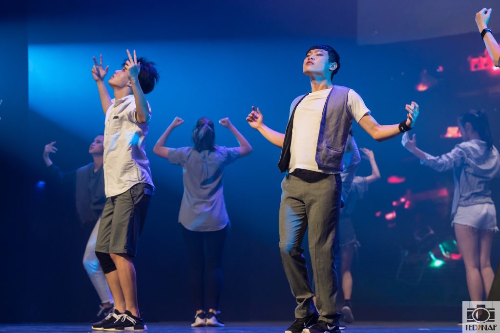 Students perform on stage under blue lighting, showcasing their dance or drama skills.