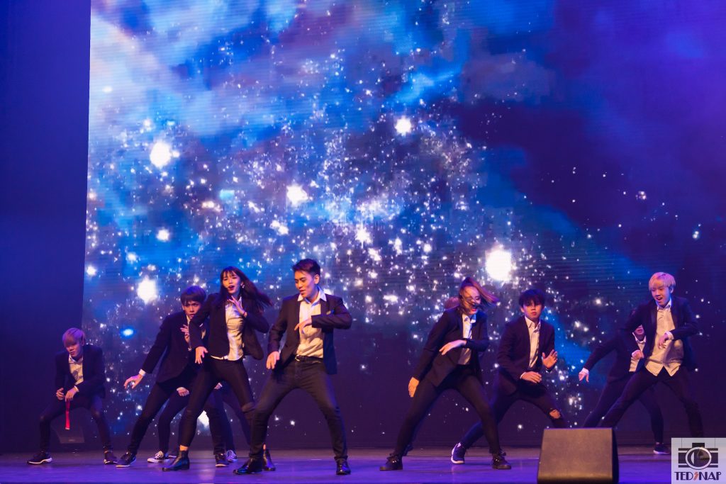 A group of performers in black suits dancing energetically on a stage with a starry, cosmic background.