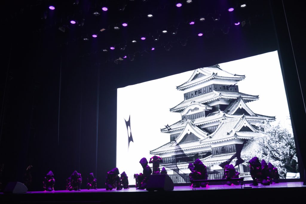 Performers on stage with a large screen displaying a Japanese castle, under vibrant purple lighting.