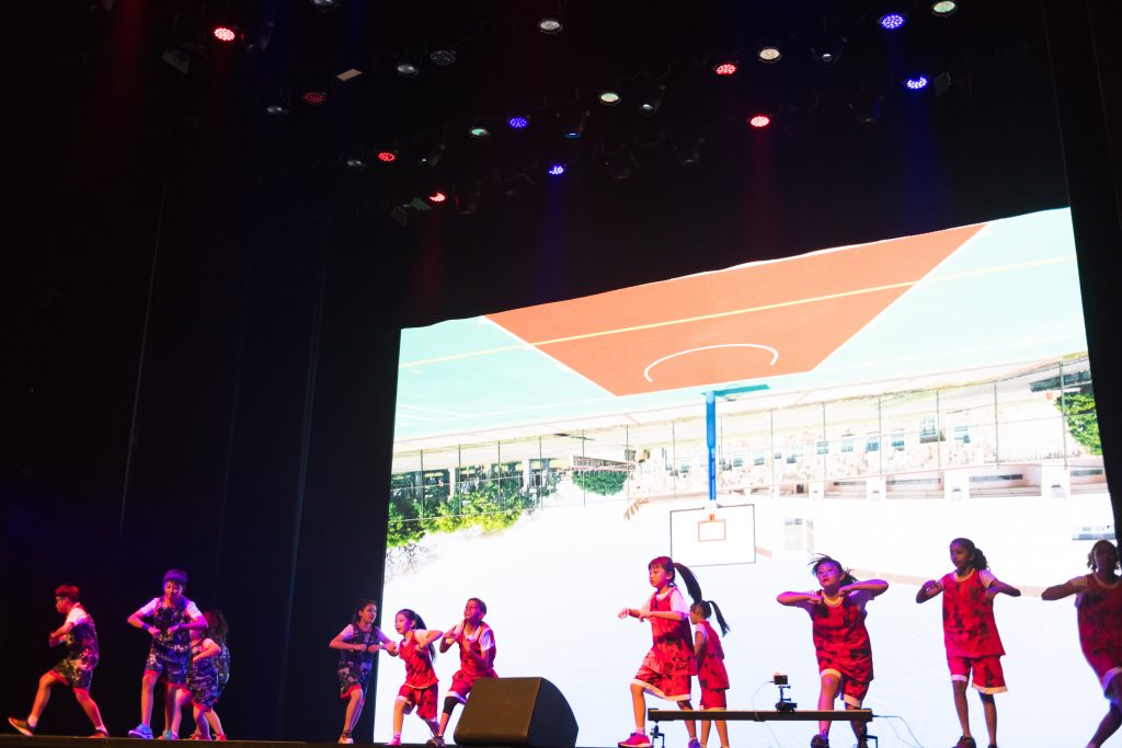 Children in sports attire perform on a stage with a basketball court projection behind them.