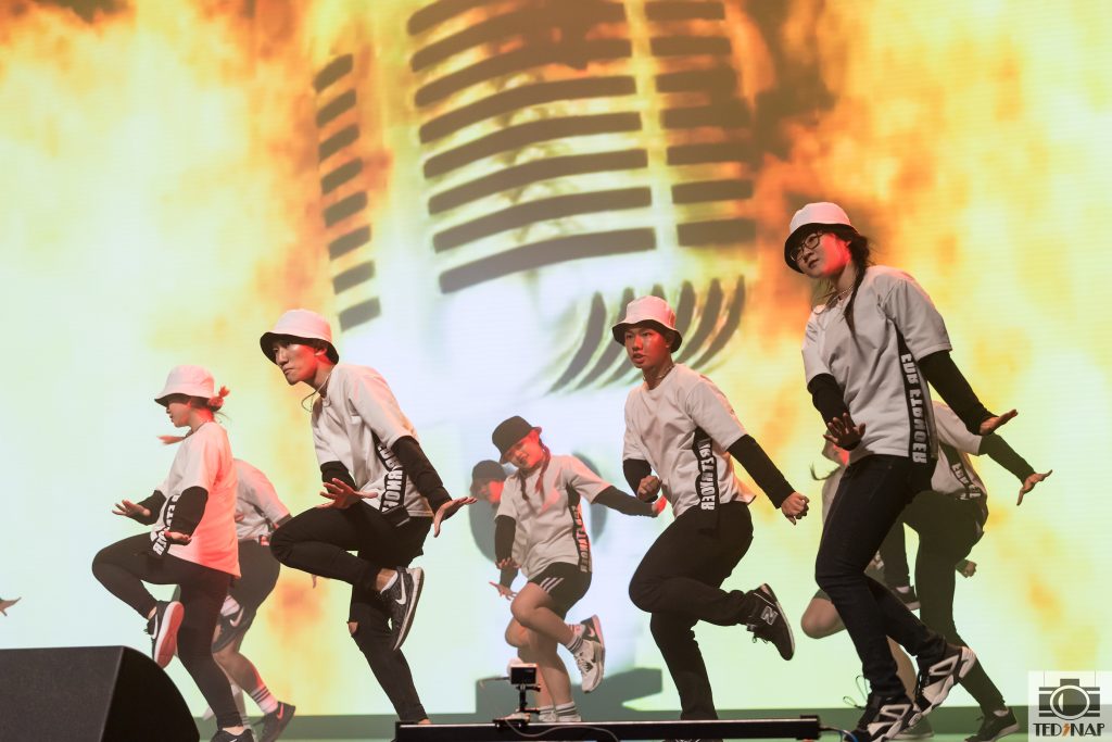 Energetic dancers in bucket hats perform on stage with a large microphone graphic backdrop.