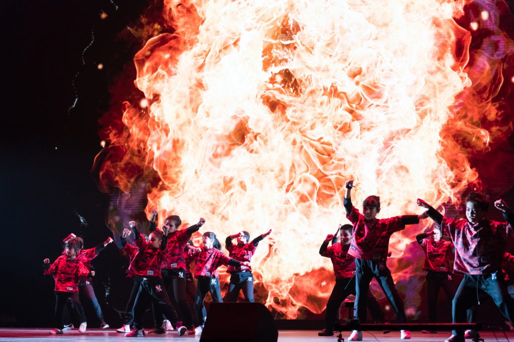Children in red and black outfits perform on stage with a dynamic fiery background display.