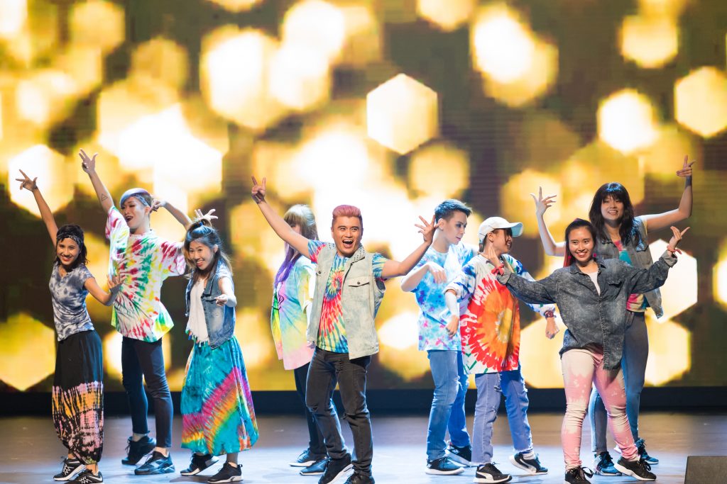 A diverse group of young people in vibrant tie-dye outfits posing energetically on a stage.