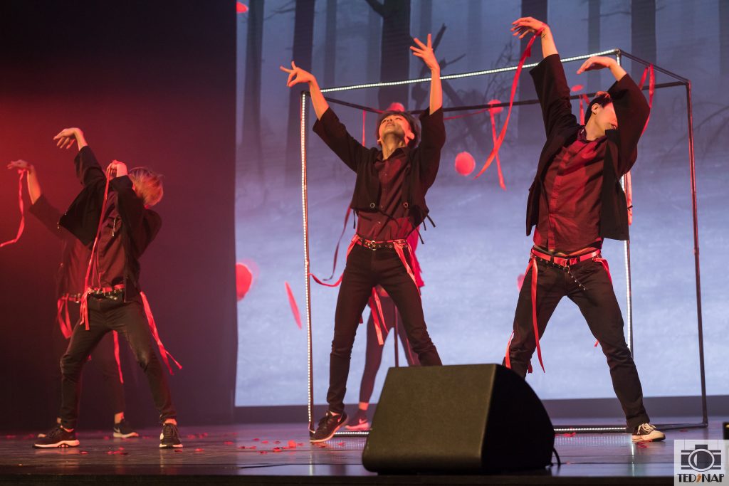 Three dancers in black and red perform on stage with a forest backdrop and falling red ribbons.