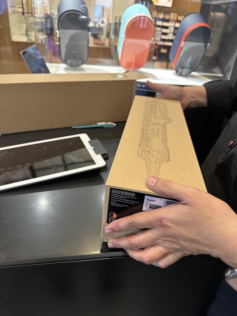 A person holds a Dyson product box, with other Dyson devices displayed in the background.
