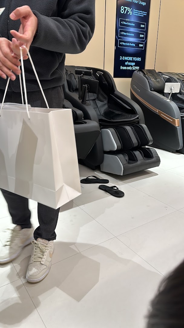 A person stands in a massage chair store, with various massage chairs and a digital display visible.