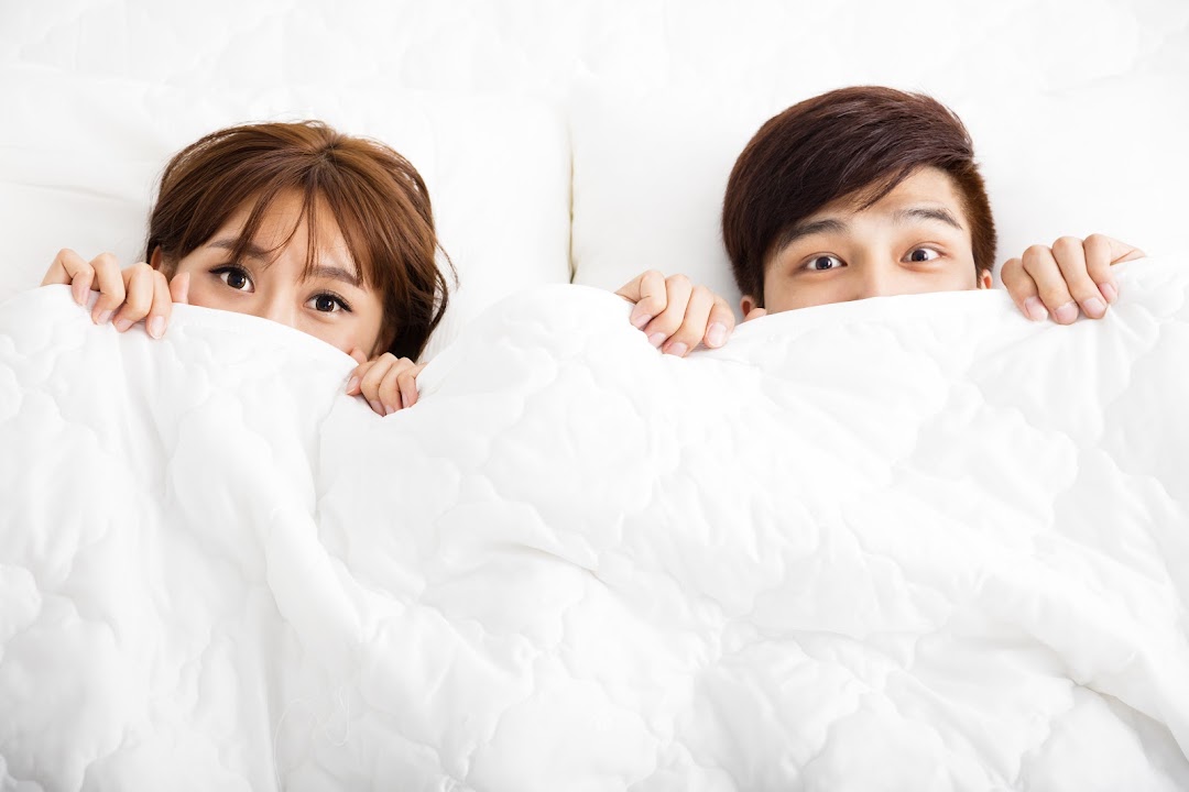 A man and a woman playfully peek from under a soft, white duvet in bed.