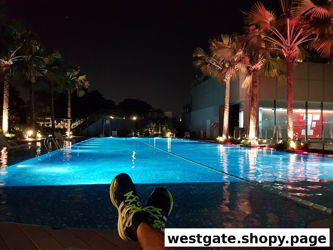 A person's feet resting by a beautifully lit swimming pool at night, surrounded by palm trees.