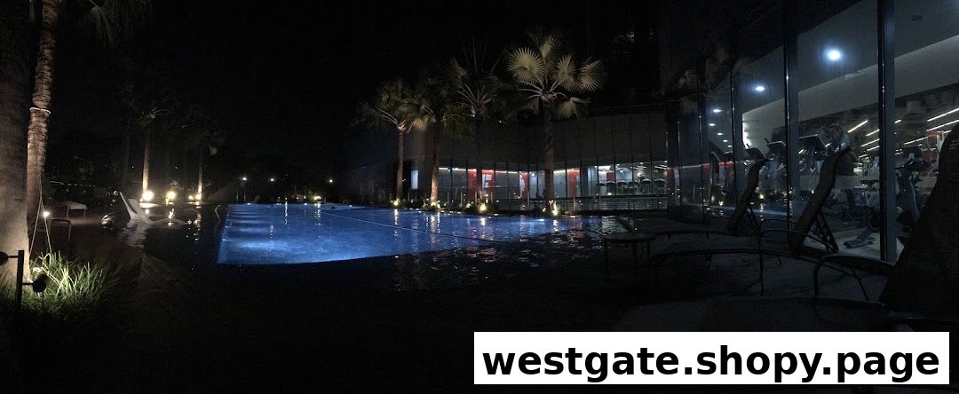 A beautifully lit swimming pool at night with a modern gym visible through glass windows in the background.
