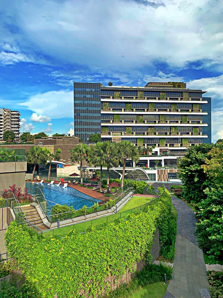 An aerial view of a modern building with a swimming pool and lush green landscaping under a blue sky.
