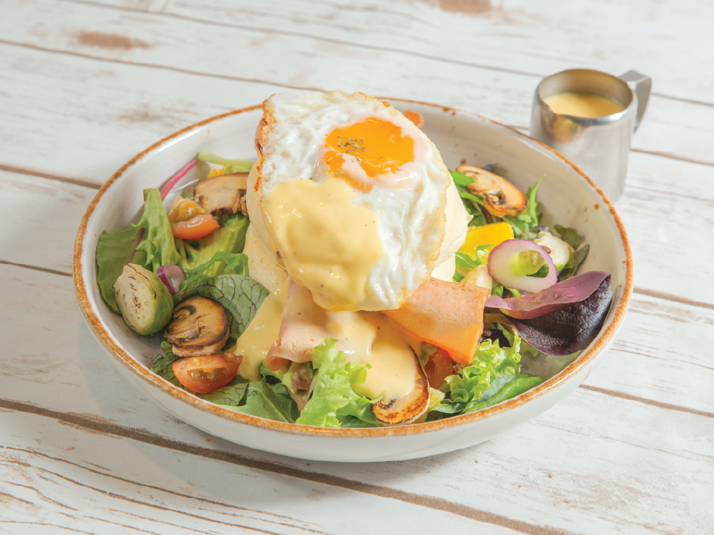 A vibrant salad bowl featuring a fried egg, hollandaise sauce, ham, and fresh greens on a rustic wooden table.