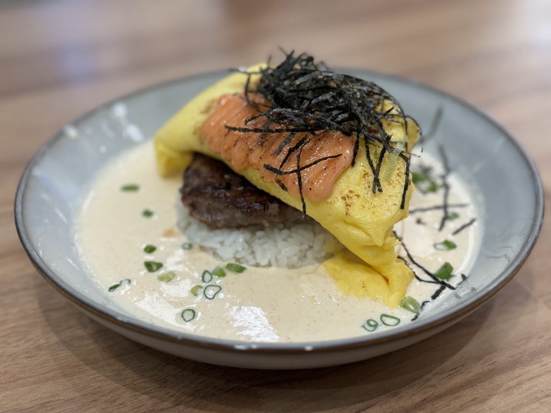 A close-up of a savory dish featuring an omelet, patty, rice, creamy sauce, and seaweed.
