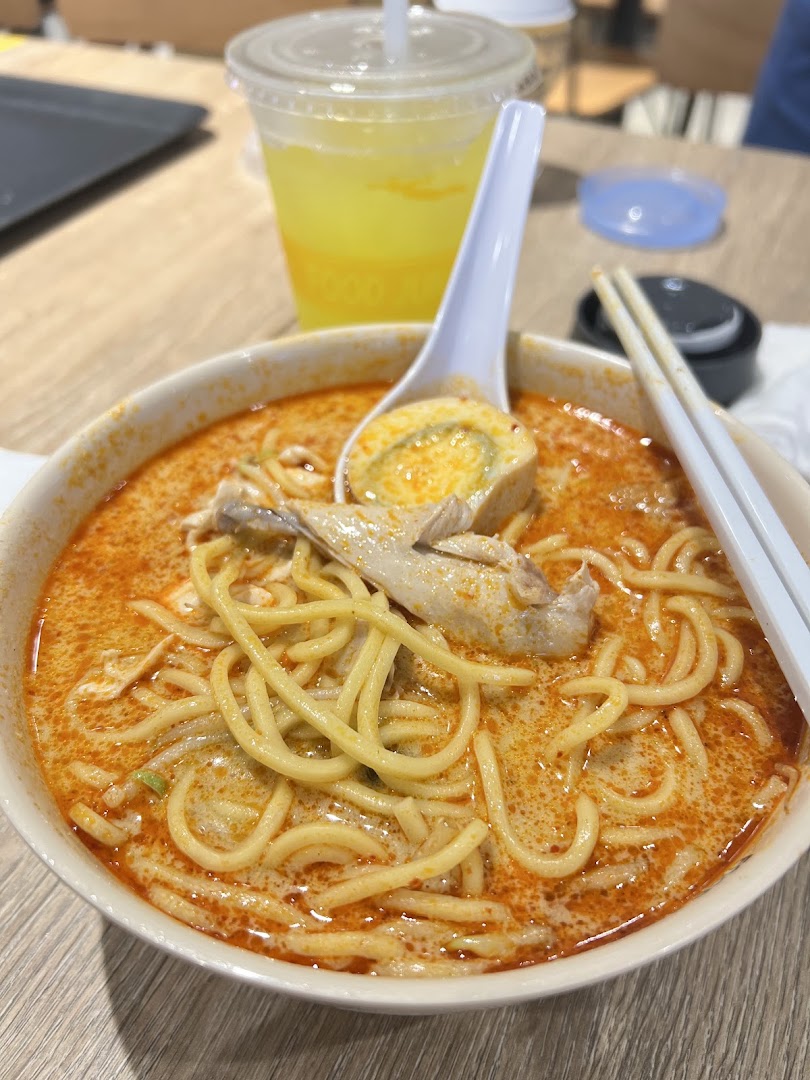 A vibrant bowl of laksa noodles with chicken and a drink on a table.