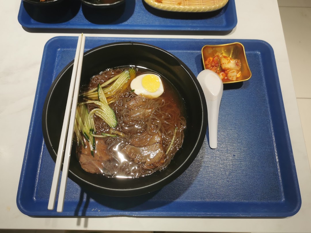A bowl of Korean cold noodles with egg, cucumber, and meat, served with kimchi.