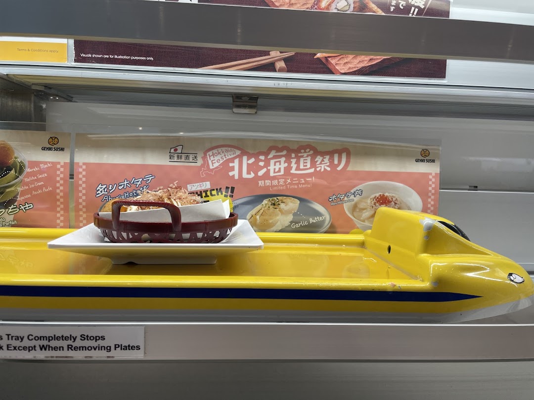 A dish on a yellow shinkansen train delivery system at Genki Sushi, with promotional posters.