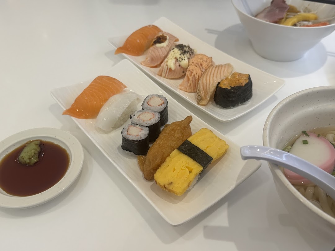 A table laden with various sushi plates, including salmon, maki, and tamago, alongside a bowl of noodles.