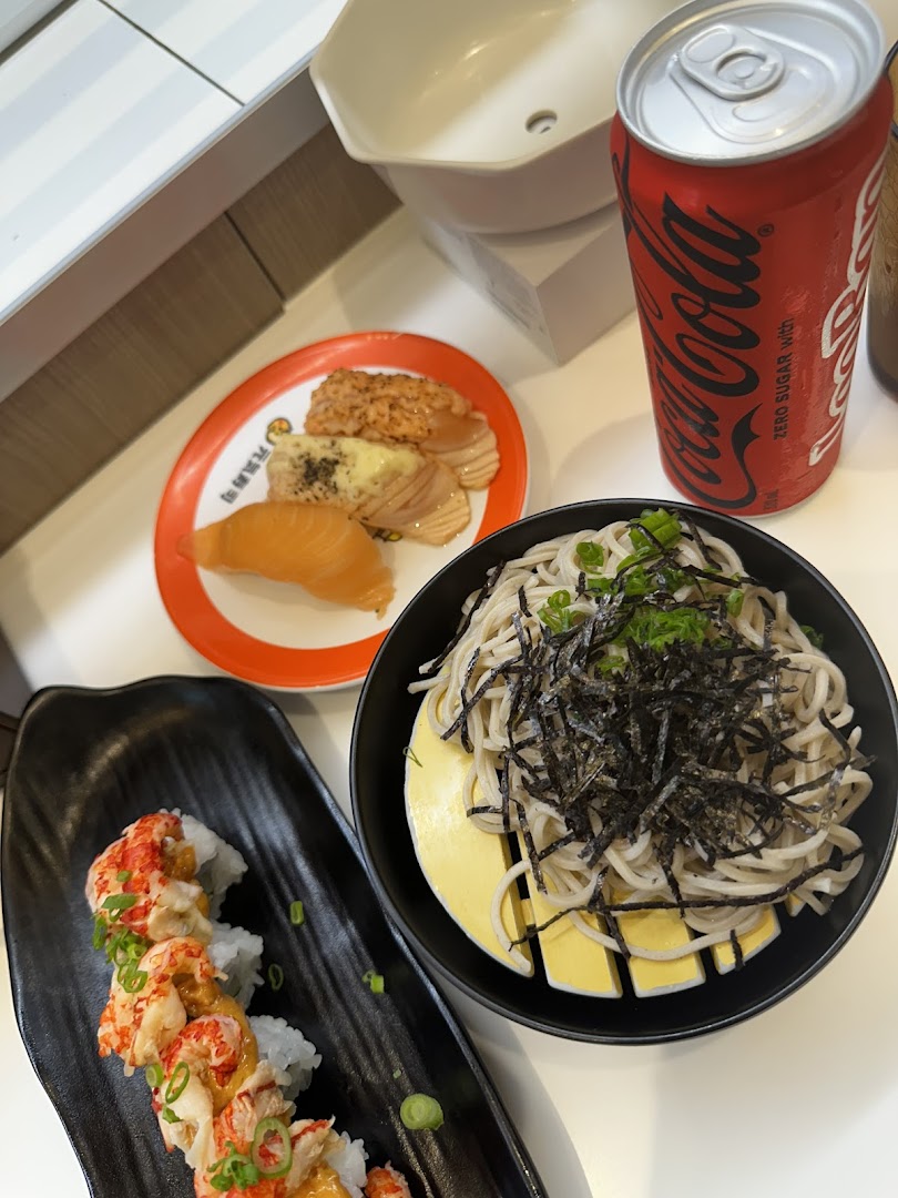 A table showcasing various sushi dishes, a noodle bowl, and a Coca-Cola can at Genki Sushi.