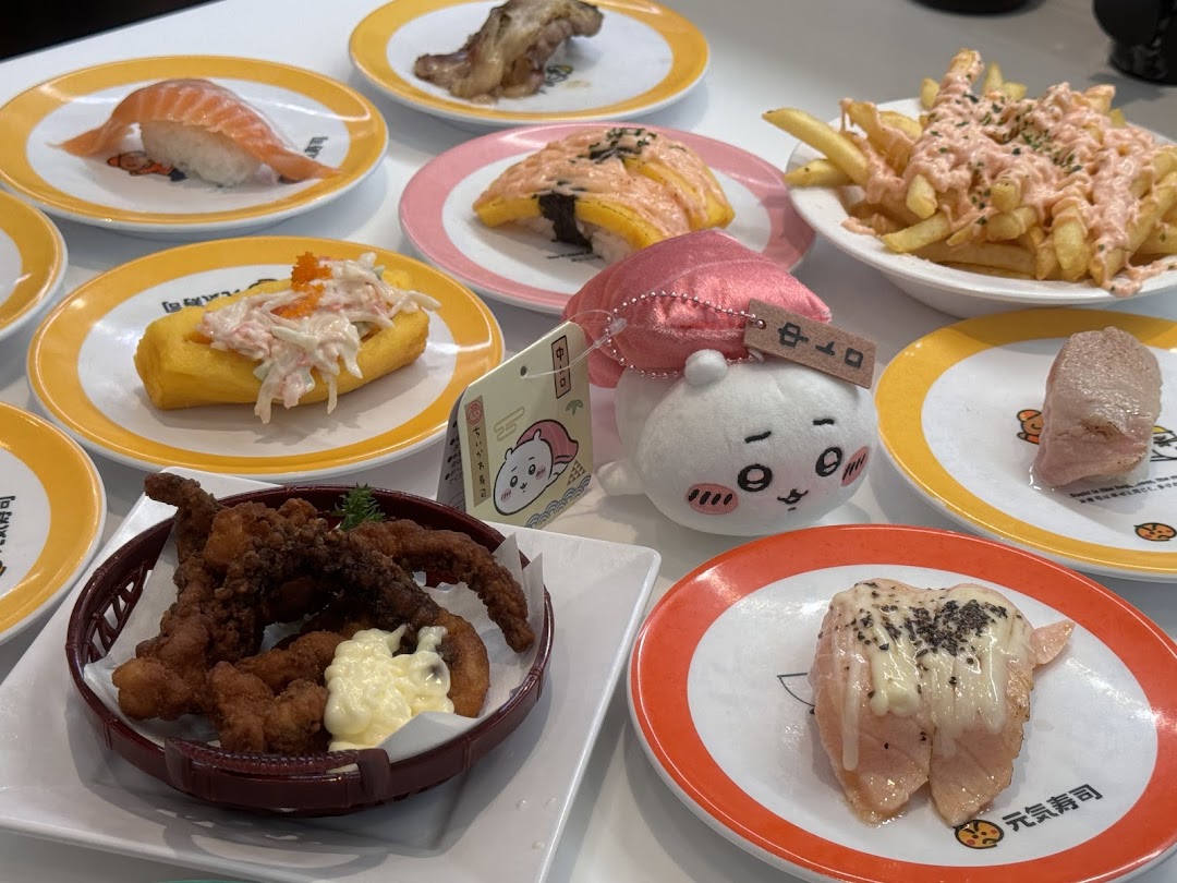 A table filled with various sushi dishes, fried food, fries, and a cute plush toy at Genki Sushi.