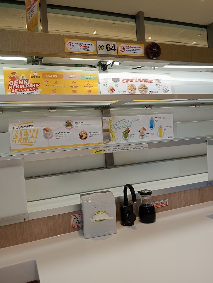 A view of a Genki Sushi counter seat with a conveyor belt and various promotional displays.