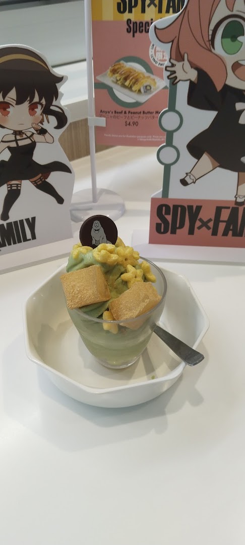 A green dessert with toppings and a spoon, alongside Spy x Family promotional standees and a small menu board.